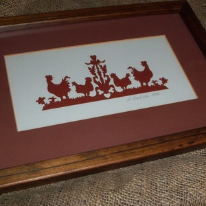 May include: A framed paper cut-out art piece depicting a group of chickens and a rooster in a red silhouette against a white background. The artwork is signed "A. Kustina, 1995".
