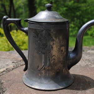 May include: A silver teapot with a floral design. The teapot has a lid with a knob on top and a spout on the side. The teapot is sitting on a gray stone surface.