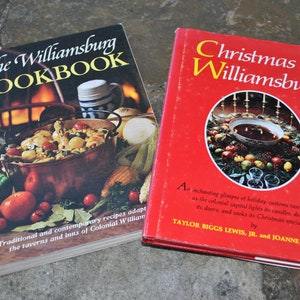 May include: Two vintage cookbooks with red and black covers. The Williamsburg Cookbook features a copper pot with food and a candle on a table. Christmas in Williamsburg features a red bowl with a Christmas wreath around it.