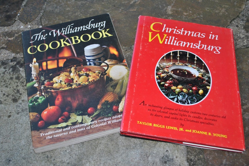 May include: Two vintage cookbooks with red and black covers. The Williamsburg Cookbook features a copper pot with food and a candle on a table. Christmas in Williamsburg features a red bowl with a Christmas wreath around it.