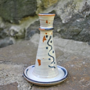 以下が含まれることがあります： 青と茶色のデザインが施された白いセラミック製のキャンドルホルダー。ホルダーは幅広のベースと、高く狭いトップを持っています。