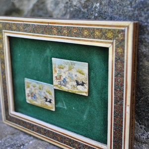 May include: Two framed miniature paintings depicting scenes of horseback riding. The paintings are set against a green velvet background and framed in a decorative ivory and wood frame with intricate mosaic details.