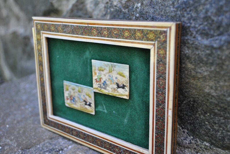 May include: Two framed miniature paintings depicting scenes of horseback riding. The paintings are set against a green velvet background and framed in a decorative ivory and wood frame with intricate mosaic details.