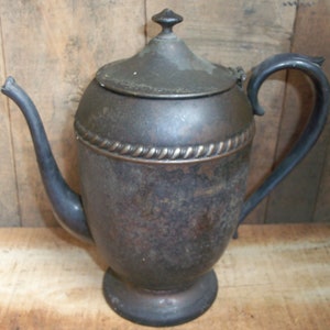 Può includere: Caffettiera antica color argento con corpo arrotondato, beccuccio e manico. Il coperchio è leggermente a cupola e ha un piccolo pomello. Un motivo decorativo a forma di corda circonda la caffettiera vicino alla parte superiore.