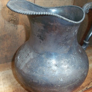 May include: A silver pitcher with a beaded rim and a decorative handle. The pitcher has a worn, aged appearance and a design etched into the side.