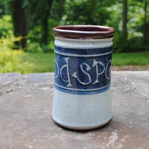 Puede incluir: Una taza de cerámica blanca con una banda azul y las palabras "Good Spot" en letras azules. La taza tiene un borde marrón.