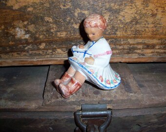 Puede incluir: Una figurita de porcelana de una niña que lleva un vestido blanco con detalles florales azules y rosas. Está sentada con las piernas cruzadas y las manos juntas en su regazo.