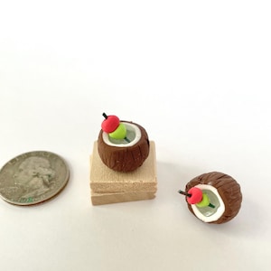 May include: Miniature coconut cocktail with a red and green fruit garnish on a wooden block. The brown coconut shell is filled with a clear liquid. A quarter is next to the cocktail for size comparison.