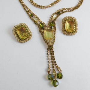 Może przedstawiać: Złoty naszyjnik z zielonym i żółtym wisiorem z cyrkonii i pasującymi kolczykami. Naszyjnik ma frędzle z zielonych cyrkonii.