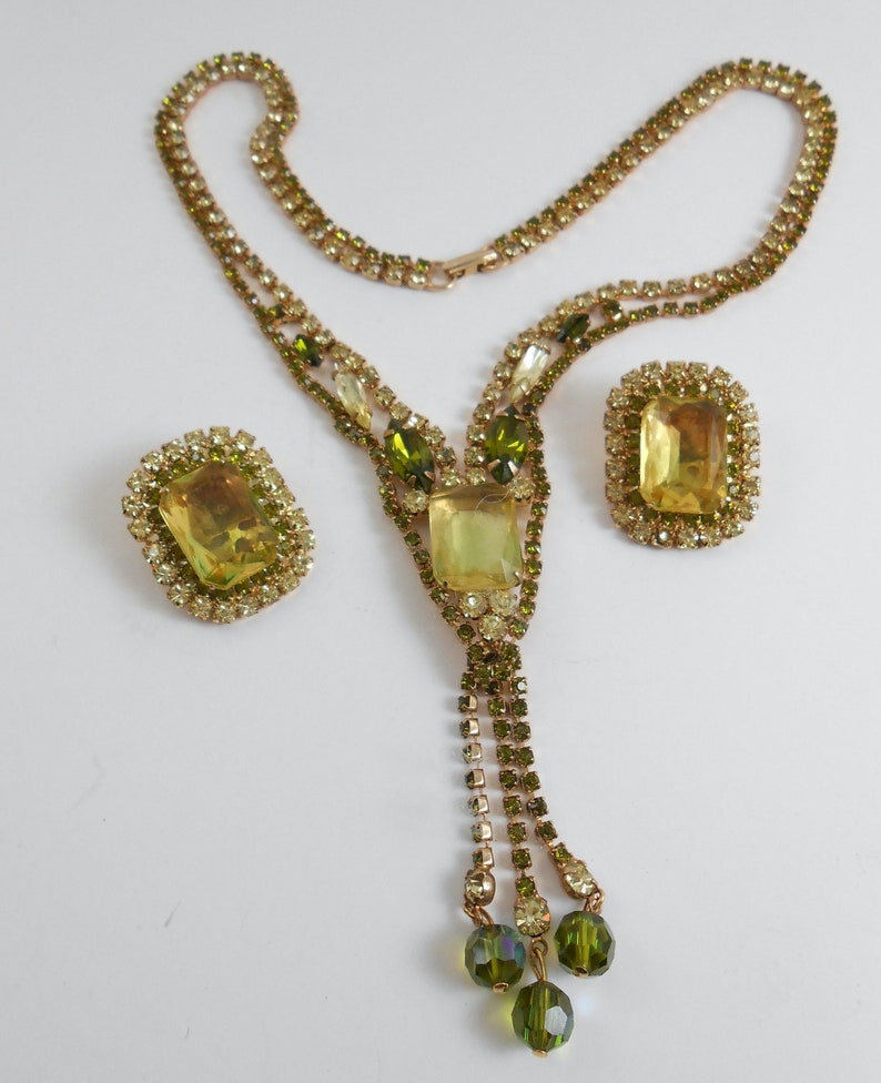 May include: A gold tone necklace with a green and yellow rhinestone pendant and matching earrings. The necklace has a tassel of green rhinestones.