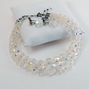 May include: A vintage clear crystal necklace and matching clip-on earrings set. The necklace features multiple strands of faceted beads, with a silver-tone clasp. The earrings have a similar crystal design. The jewelry set is displayed on a white surface.