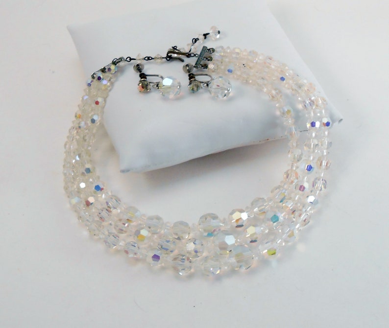 May include: A vintage clear crystal necklace and matching clip-on earrings set. The necklace features multiple strands of faceted beads, with a silver-tone clasp. The earrings have a similar crystal design. The jewelry set is displayed on a white surface.