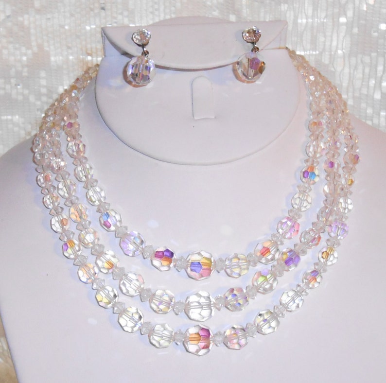 May include: A clear, iridescent crystal necklace and matching earrings set. The necklace features three strands of faceted beads, with larger, hexagonal beads interspersed. The earrings are small, round crystal studs. The jewelry is displayed on a white mannequin.
