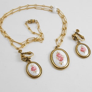 May include: A gold chain necklace with a white oval pendant featuring a pink rose design. Matching clip-on earrings with the same design.