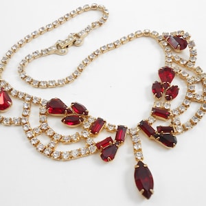 Puede incluir: Un collar de tono dorado con un diseño de pedrería roja y transparente. El collar presenta un diseño en cascada con un colgante en forma de lágrima.