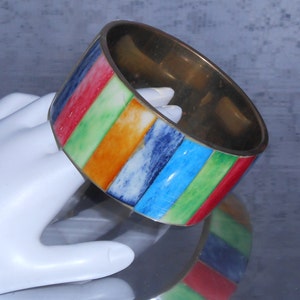 May include: A brass bangle bracelet with a colorful striped design. The stripes are in red, green, orange, blue, and white. The bracelet has a polished, metallic finish and a smooth interior.