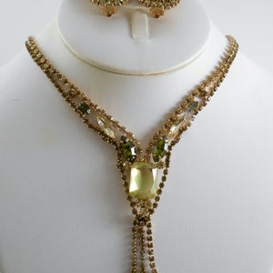 May include: A gold tone necklace and earrings set with a large, light green, rectangular rhinestone in the center of the necklace and smaller green rhinestones surrounding it. The necklace has a tassel of smaller rhinestones.