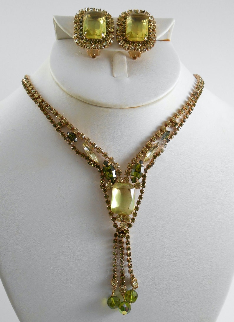 May include: A gold tone necklace and earrings set with a large, light green, rectangular rhinestone in the center of the necklace and smaller green rhinestones surrounding it. The necklace has a tassel of smaller rhinestones.