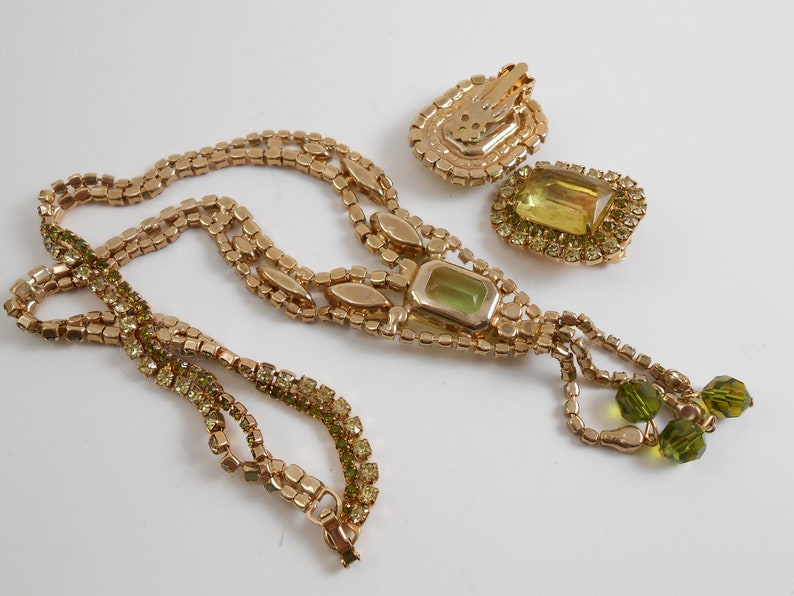 May include: A gold tone necklace and earrings set with a large green stone and smaller green rhinestones. The necklace has a tassel of green beads.
