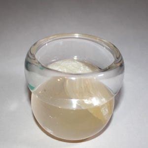Könnte beinhalten: Ein klarer, transparenter Ring mit einer abgerundeten, bauchigen Basis. Das Band des Rings ist dick und glatt, und die Basis hat eine helle, durchscheinende Farbe. Der Ring scheint aus Glas oder einem ähnlichen Material zu bestehen.