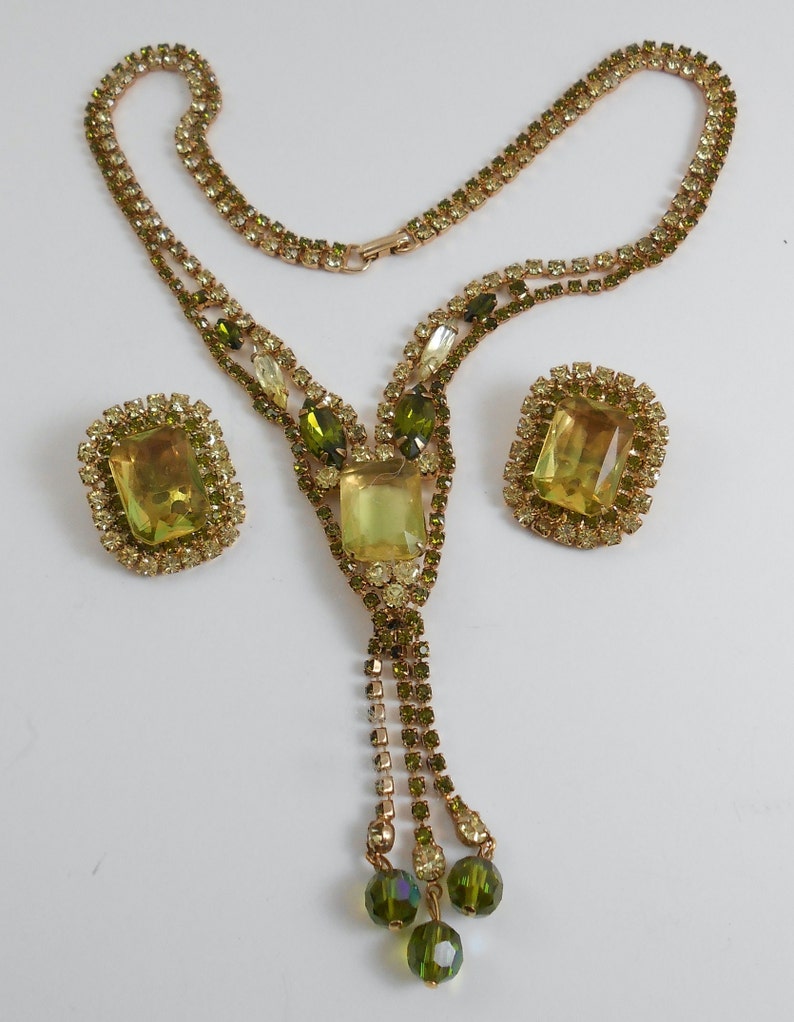 May include: A vintage green and yellow rhinestone necklace and earrings set. The necklace features a long, dangling tassel with three green beads at the end. The earrings are rectangular with a large, faceted, yellow rhinestone in the center.
