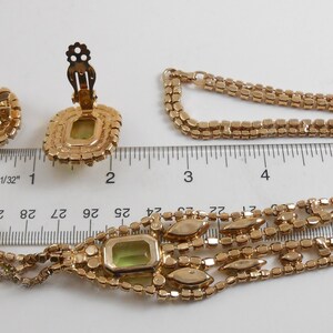 May include: A gold tone necklace and earrings set with a green rhinestone pendant and matching earrings. The necklace is a multi-strand chain with a drop design. The pendant is a square shape with a green rhinestone in the center. The earrings are clip-on style and have a square shape with a green rhinestone in the center.