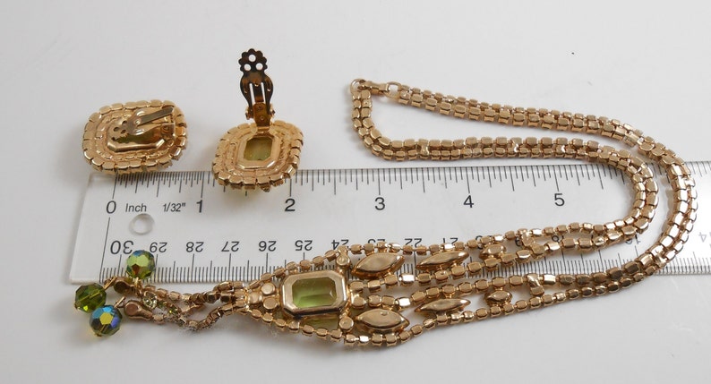 May include: A gold tone necklace and earrings set with a green rhinestone pendant and matching earrings. The necklace is a multi-strand chain with a drop design. The pendant is a square shape with a green rhinestone in the center. The earrings are clip-on style and have a square shape with a green rhinestone in the center.