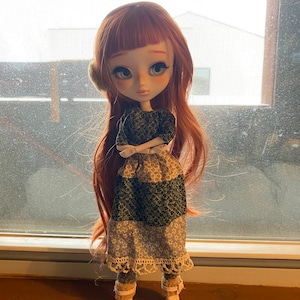 May include: A red-haired doll with green eyes wearing a green and white dress, white boots, and white earmuffs. The doll is standing in front of a window.