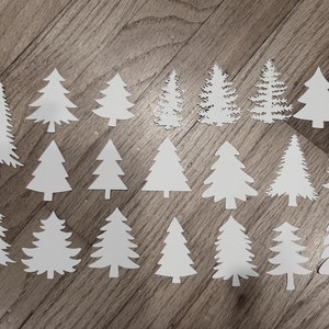 Puede incluir: Una colecci&oacute;n de recortes de &aacute;rboles de Navidad de papel blanco en varios estilos y tama&ntilde;os, dispuestos sobre una superficie de madera. Los &aacute;rboles var&iacute;an desde formas triangulares simples hasta dise&ntilde;os detallados y en capas, ideales para manualidades y decoraciones navide&ntilde;as.