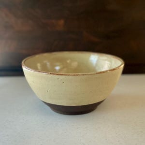 May include: A ceramic bowl featuring a two-tone design with a creamy off-white glazed upper section and a dark brown unglazed base. The rim is thin and rounded, showing a subtle brown edge that complements the dark bottom of the bowl.