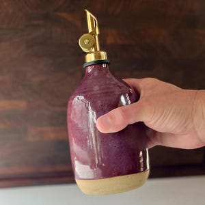May include: A ceramic oil dispenser with a glossy, deep purple glaze on the upper body and a raw, unglazed beige base. The vessel features a metallic gold-toned pour spout with a hinged lid mechanism, providing a functional and sleek design.