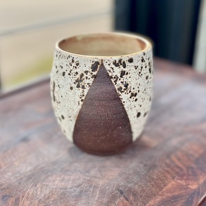 May include: A ceramic tumbler with a speckled white exterior and a dark brown, triangular accent. The interior is a light beige. The cup is designed with a rounded base and a wide opening, suitable for beverages.