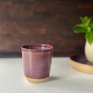 May include: A handmade ceramic tumbler with a deep purple glaze and a natural beige base. The cup has a smooth, glossy finish and a simple, cylindrical shape. The background is a dark wood surface.