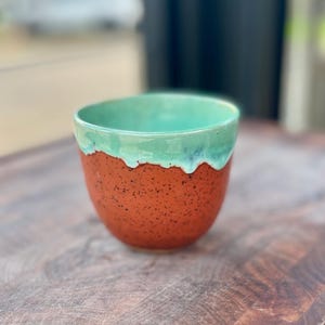 Könnte beinhalten: Eine handgefertigte Keramiktasse mit gesprenkeltem orangefarbenem Boden und türkisfarbenem glasiertem Rand. Die Tasse hat eine abgerundete Form und eine glatte, glänzende Oberfläche. Der Innenraum ist ebenfalls türkis. Die Tasse steht auf einer Holzoberfläche.
