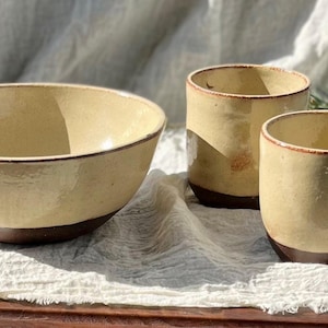 May include: A ceramic bowl and two matching cups. The bowl is large and round, with a light yellow interior and a dark brown base. The cups are cylindrical, also with a light yellow exterior and dark brown bases. The set is placed on a white cloth.