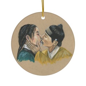 Pode incluir: Um ornamento redondo de cerâmica com uma ilustração pintada à mão de duas pessoas se beijando. A mulher está vestindo uma roupa verde e azul, e o homem está vestindo uma roupa amarela e dourada. O fundo é marrom claro.