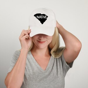 May include: White baseball cap with a black outline of the state of South Carolina and the word "NATIVE" in white text.