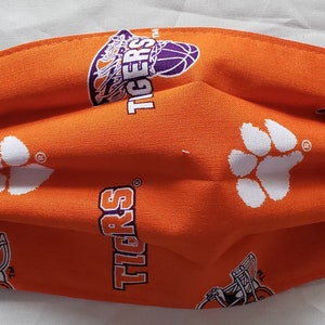 May include: Orange face mask with a white paw print and the word "Tigers" repeated on the fabric. The mask has purple elastic straps.