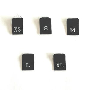 May include: Five black fabric labels with white text indicating sizes: XS, S, M, L, and XL. The labels are rectangular with rounded corners and are arranged vertically. The background is plain white.