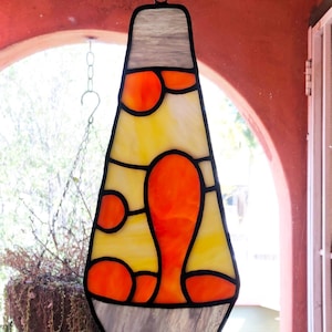 May include: A stained glass lava lamp hanging in a window. The lamp is yellow and orange with a gray base.
