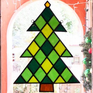 May include: A stained glass Christmas tree ornament with a diamond pattern in shades of green and yellow. The ornament is hanging in a window with a red frame.