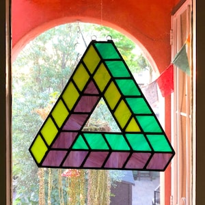 May include: A stained glass window hanging in a window frame. The window is shaped like a triangle and features a geometric pattern of yellow, green, and purple glass.