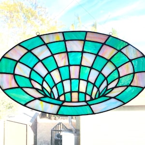 Op de afbeelding: Een gebrandschilderd raam dat in een raamkozijn hangt. Het gebrandschilderde glas is trechtervormig en gemaakt van groen, roze en wit glas.