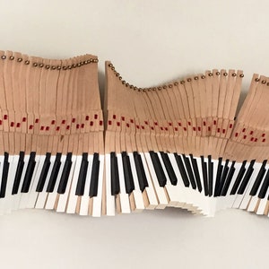 May include: A unique wall art piece featuring a piano keyboard design. The artwork is composed of individual keys and hammers, with black and white keys and tan-colored hammers. The piece has a wavy, abstract form and is mounted on a white wall.