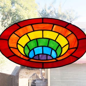 May include: Oval stained glass window panel with a rainbow design. The panel has a red border, with concentric rings of orange, yellow, green, blue, and purple. The glass is divided into geometric shapes by black lines.