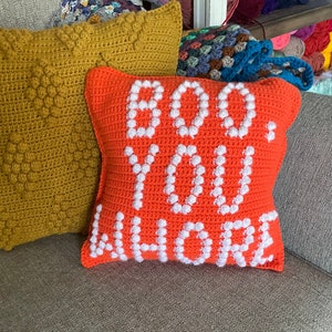 May include: Two crocheted pillows, one orange with white text that says "BOO YOU WHERE" and one mustard yellow with a textured pattern.