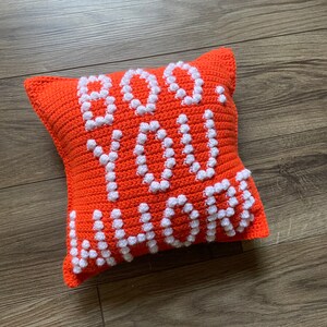 May include: Orange crocheted pillow with white bobbles spelling out "BOO YOU" in a playful font.