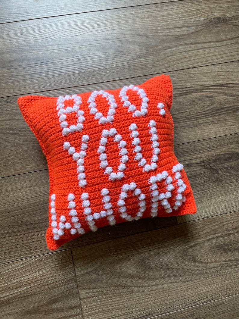 May include: Orange crocheted pillow with white bobbles spelling out "BOO YOU" in a playful font.