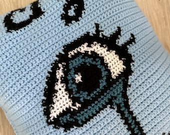 May include: A light blue crocheted pillow with a black and white eye design. The eye is outlined in black and has a blue iris and a black pupil.