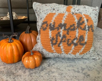 May include: A crocheted pillow with a pumpkin design and the text "Pumpkin Spice" in brown lettering. The pillow is on a gray carpet with three orange pumpkins.